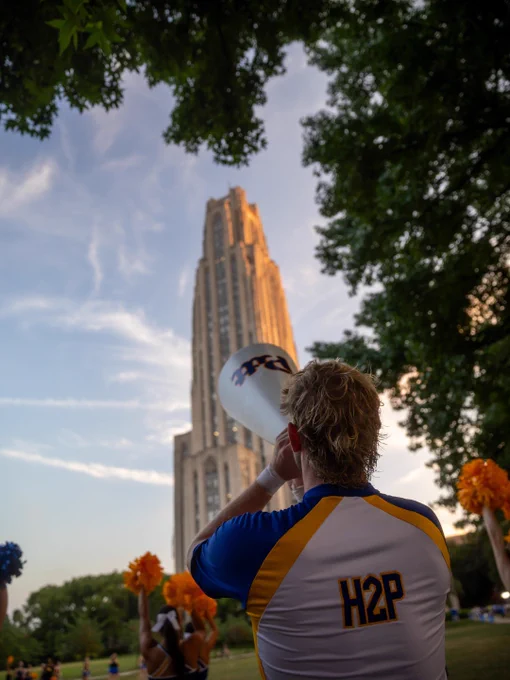 A person wearing a shirt with "H2P" on the back holds a megaphone and stands in front of the Cathedral of Learning at the University of Pittsburgh. Nearby, people are holding orange pom-poms. Trees frame the top and sides of the image.