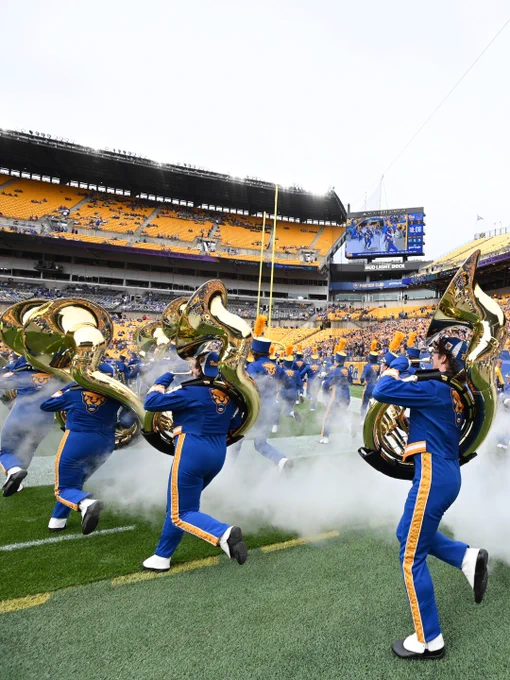Pitt marching band performs energetically at a football stadium, with members playing sousaphones in vibrant blue and gold uniforms. Smoke creates a dynamic effect around the band. The venue is Acrisure Stadium, with a large screen and scattered spectators visible in the stands.