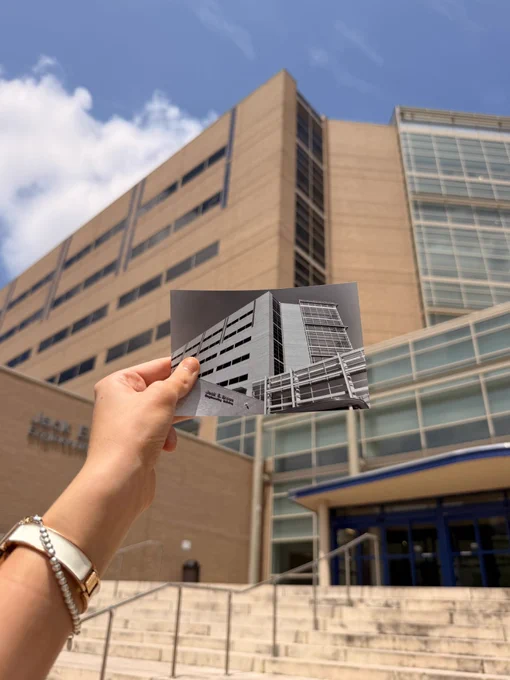A hand holds a black and white photo of the Chase Tower building against the same building in the background, highlighting the architectural similarities. The sky is clear with a few clouds.