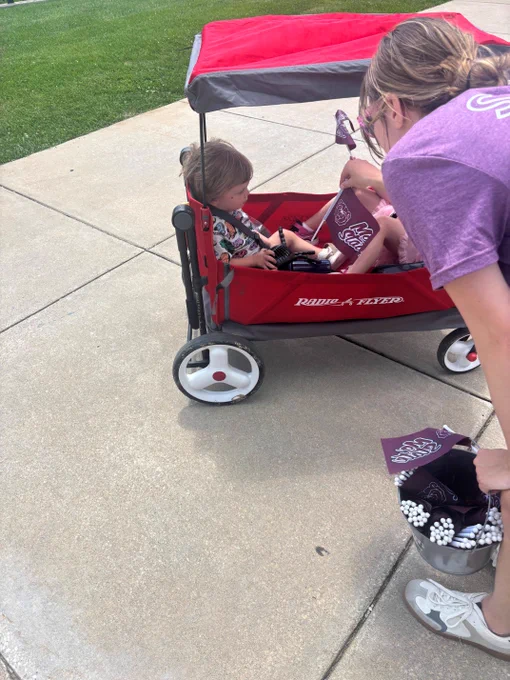 A person is pushing a red Radio Flyer wagon with a child sitting inside. The child is holding a small Megabucks toy. The person is wearing purple attire and sneakers, and is holding pom-poms. The scene is outdoors on a concrete sidewalk with grass visible on the side.