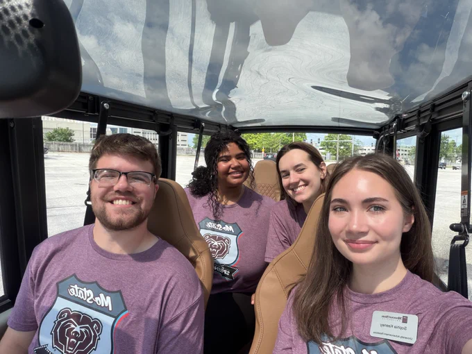 A group of four people sitting in an open-top vehicle, all wearing matching t-shirts featuring a design with 'Mo State' and a bear logo. They are smiling, and sunlight casts shadows on the vehicle's transparent roof.