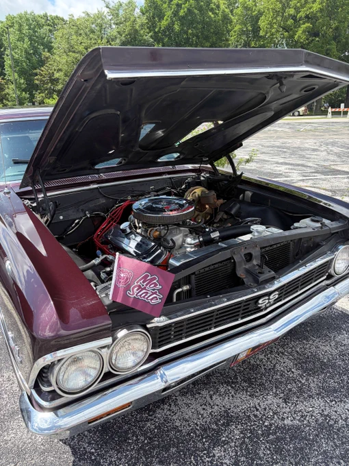 A classic car with an open hood revealing its engine. The car has a maroon exterior, and a sign stating “No Subs” is displayed inside the engine compartment. The car is parked on a paved surface with greenery visible in the background.