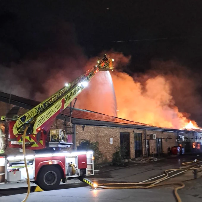 A turntable ladder is parked in front of a brick warehouse. Firefighters are spraying water from the extended ladder, onto the burning warehouse