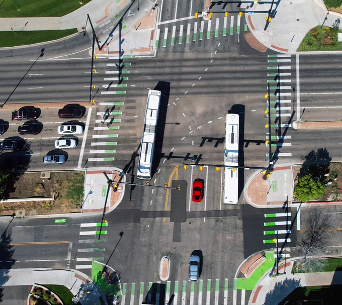 A bird's eye view of an intersection with bike lanes and crosswalks. Bus and vehicle traffic travel through the roads.