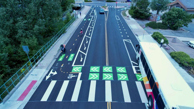 A bird's eye view of a crossing with a bus, a car and two bicycles traveling along the road.