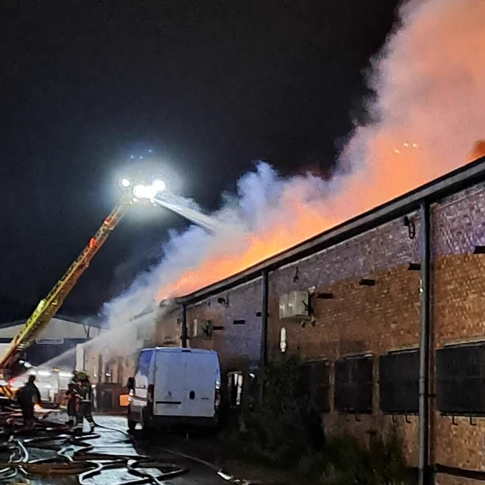A turntable ladder is parked in front of a brick warehouse. Firefighters are spraying water from the extended ladder, onto the burning warehouse. A white van is also parked against the side of the building