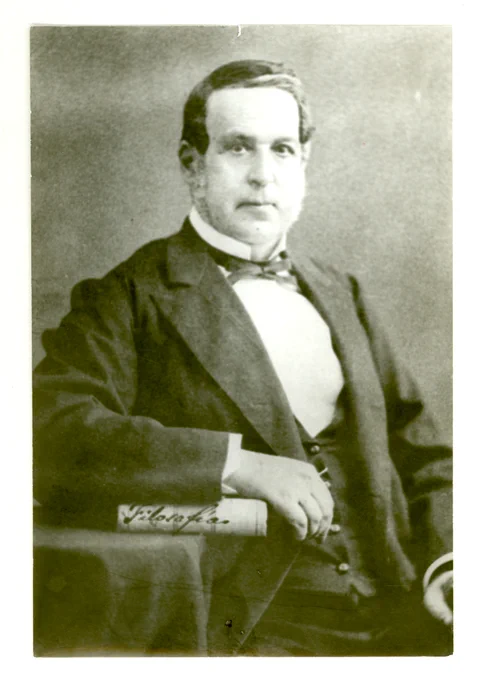 Retrato de Ramón Briseño en blanco y negro. Hombre vestido con traje formal y pajarita, apoyando un brazo sobre una mesa con un libro.