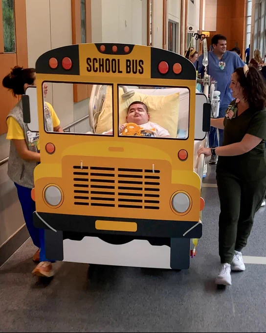 A person in a hospital bed is being transported by two individuals, with the bed decorated to resemble a school bus. The scene takes place in a hospital corridor.