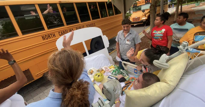 A group of people are gathered outside near two North Carolina public school buses. One person is lying in a hospital bed with supportive medical equipment. Others around the bed are waving and interacting with those on the bus.