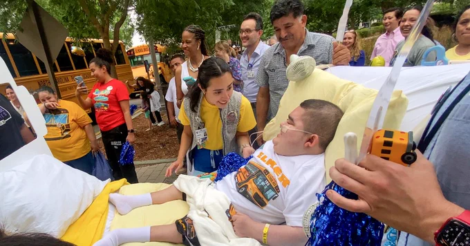 A group of people are gathered outside near two North Carolina public school buses. One person is lying in a hospital bed with supportive medical equipment and another person is talking with them. 