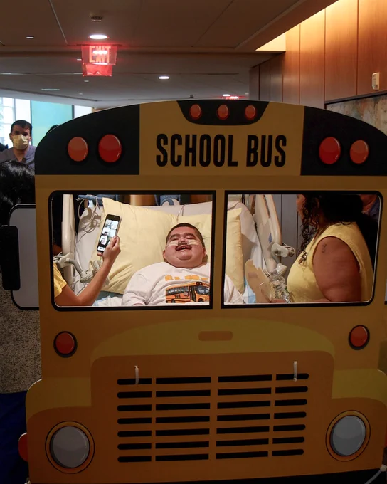 A person in a hospital bed, surrounded by others, is smiling while framed by a school bus cutout. A phone is held up by one of the people around them.