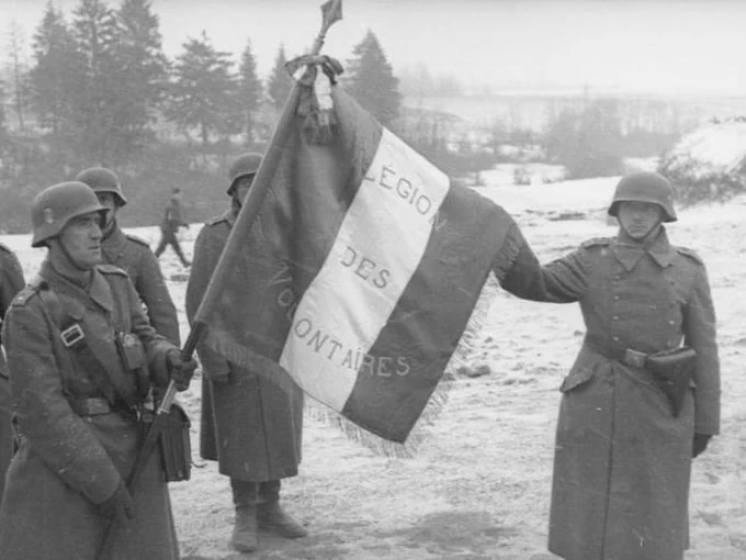 A group of soldiers in winter uniforms stands in a snowy landscape. One soldier holds a flag with the inscription "Légion des Volontaires." Trees and a few structures are visible in the background.