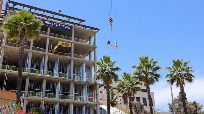 A multi-story building under construction with a visible Hensel Phelps banner. A crane is positioned above, and several palm trees surround the site, with a clear blue sky in the background.