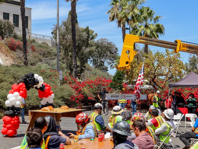 A group of construction workers in safety vests and helmets sit at a table outdoors with pizza and drinks. Nearby, a crane holds an American flag. Black, white, and red balloons form an arch, and there are Coca-Cola cans on the table. Trees and buildings are visible in the background.