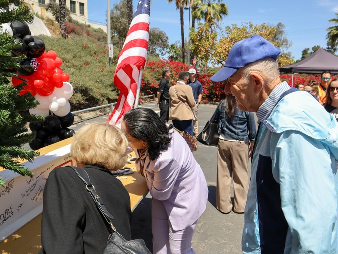 A person in a purple outfit signs a memorial board covered with messages at an outdoor event. Another person watches nearby. An American flag and a balloon arrangement in red, black, and white are in the background. People are gathered around, and the setting includes trees and a tent.