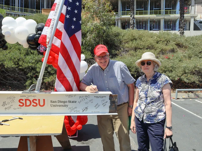 Two people stand beside a signed steel beam at a San Diego State University event. The beam is adorned with numerous signatures. One person is signing the beam while wearing a red "Aztecs" cap. An American flag and a cluster of balloons are in the background.
