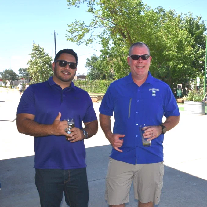 Two TAMU-CC Alumni standing outdoors on a sunny day, wearing sunglasses and holding drinks. Both wear casual blue shirts. Greenery and trees are visible in the background.