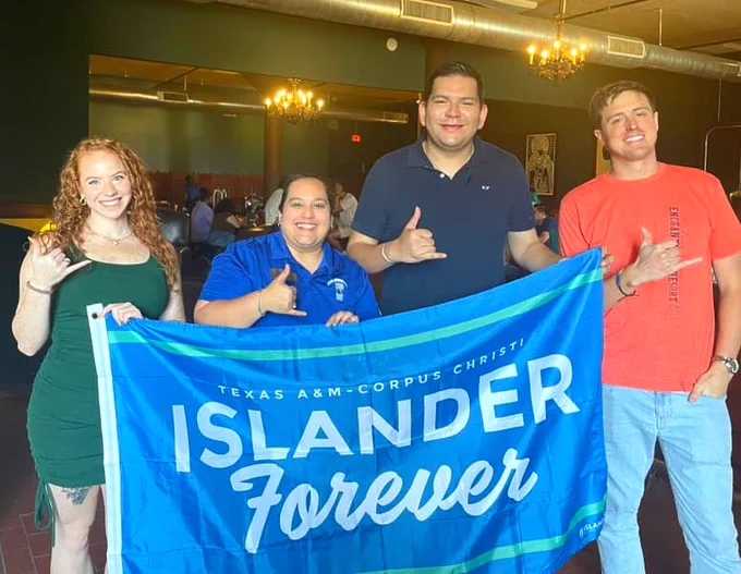 A group of four TAMU-CC Islander alumni stands indoors holding a blue banner that reads "Islander Forever, Texas A&M University-Corpus Christi." They are posing and making a shakas up hand gesture, with a dining area visible in the background.