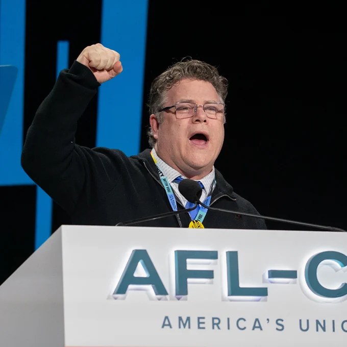 SAG-AFTRA National President Sean Astin is raising a clenched fist while speaking at an AFL-CIO podium during a conference.