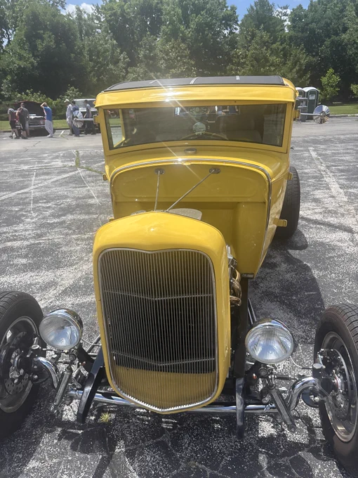 A bright yellow vintage car with a classic design is parked in a lot. The car features a prominent grille, round headlights, and polished chrome details. Trees are visible in the background.