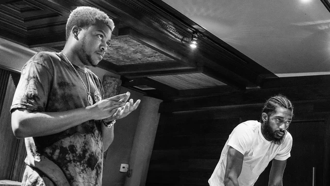 A black-and-white photo of Bino Rideaux and Nipsey Hussle in a studio, with Bino standing to the left and Nipsey leaning over a table on the right.