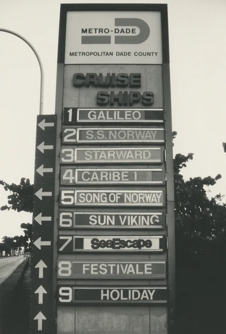 Black and white photo of a tall directory sign for \"Metro-Dade Metropolitan Dade County Cruise Ships.\" It lists nine ships: 1 Galileo, 2 S.S. Norway, 3 Starward, 4 Caribe 1, 5 Song of Norway, 6 Sun Viking, 7 SeaEscape, 8 Festivale, 9 Holiday, with directional arrows.
