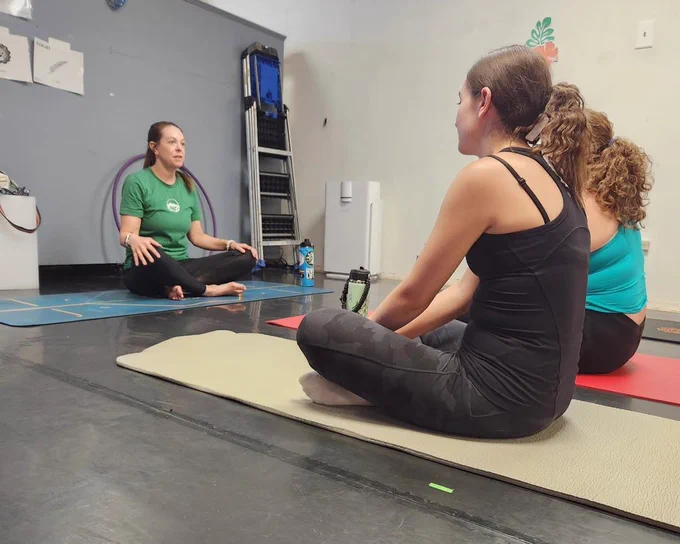 Three women sitting cross-legged on yoga mats in a studio, with one instructor guiding the session in a green shirt
