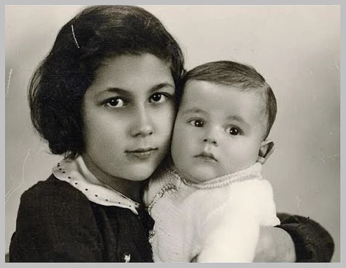Vintage black and white photograph of a young girl tenderly holding a baby, both gazing directly at the camera, against a neutral backdrop.
