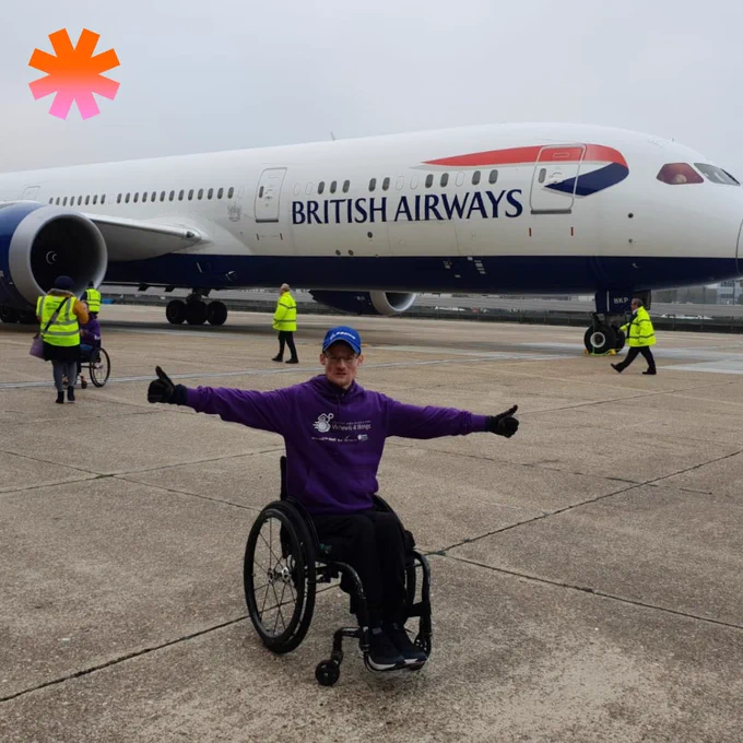 Person in a wheelchair wearing a purple hoodie and blue cap poses with arms outstretched in front of a British Airways airplane on the tarmac.