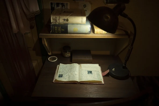 A dimly lit desk features an open diary with handwritten notes and small pasted images. Above, a shelf holds several books, including "Anne Frank: The Diary of a Young Girl." A vintage lamp illuminates the scene.