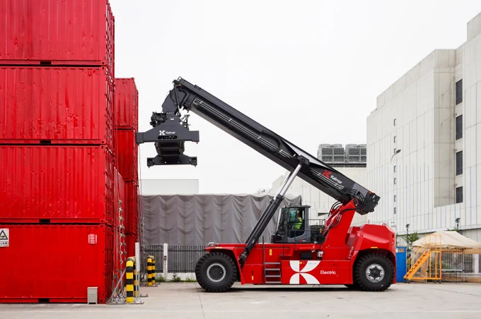 Read Kalmar Electric Reachstacker lifting a shipping container at an industrial port with stacked red containers in the background.