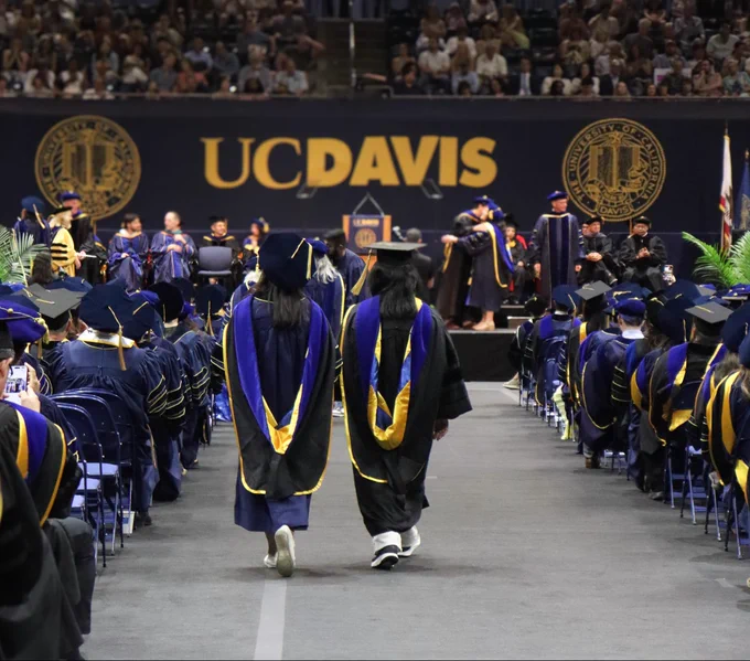 Doctoral graduate with faculty walking down the commencement aisle with the yellow UC Davis sign above them.