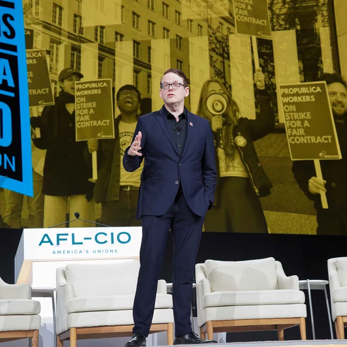 National Executive Director & Chief Negotiator Duncan Crabtree-Ireland in a dark suit speaking on stage with AFL-CIO backdrop showing union strike signs.