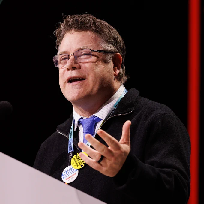 SAG-AFTRA President Sean Astin is wearing a black jacket and blue tie speaking at a podium with badges on their lanyard against a dark background.