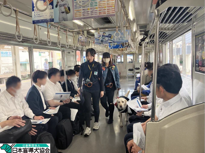 電車の車内で、通路を協会職員が盲導犬ユーザーを誘導している様子。周囲の座席には駅従業員が並んで座っており、誘導の様子を見ている。
