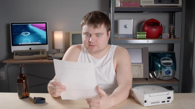 A disheveled Krazy Ken sitting at the desk in his lair. His hair is messy and face is unshaven, while wearing a white tank top. A Sega Dreamcast and empty brown bottle are sitting next to him while he stares at a piece of paper in his hand with a shocked face.