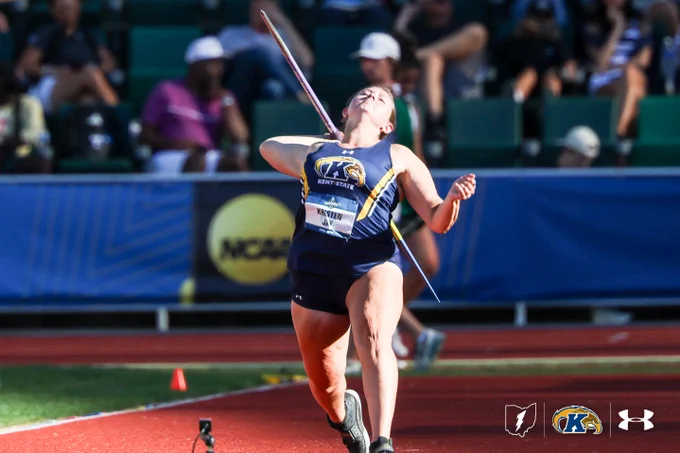 An athlete from Kent State University is in mid-action throwing a javelin at an outdoor track and field event. The background shows spectators in a stadium. Logos for NCAA and Under Armour are visible.