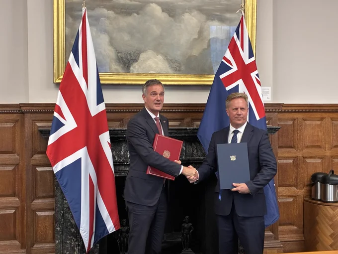 New Zealand Minister for Trade and Investment Todd McClay shakes hands with his UK counterpart, Secretary of State Peter Kyle. Behind them are the respective flags of their countries.