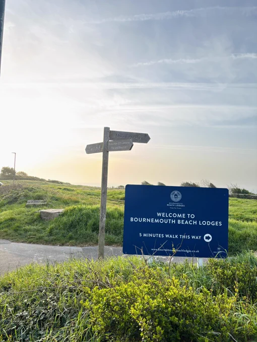 A finger post sign next to a large sign, surrounded by a grass area under a dawn sky