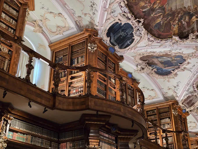 Zu sehen ist ein Teil der prächtigen Stiftsbibliothek St. Gallen.
Das Foto wurde von unten aufgenommen. Es zeigt von unten eine Galerie mit einem geschwungenen Verlauf und einem reich verzierten Holzgeländer. Einige Bücherregale, die komplett gefüllt sind, sind ebenfalls zu sehen. Ein Teil des Deckenfreskos ist ebenfalls zu sehen.