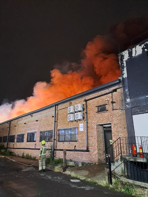 Night time photo showing a firefighter stood in front of a brick warehouse. Smoke can be seen rising from the building, coloured red by the glow of the flames. 