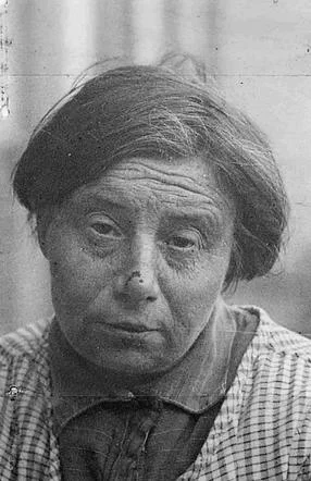 Close-up portrait of an elderly woman with a solemn expression, wearing a plaid shirt and a dark headband, set against a blurred background.