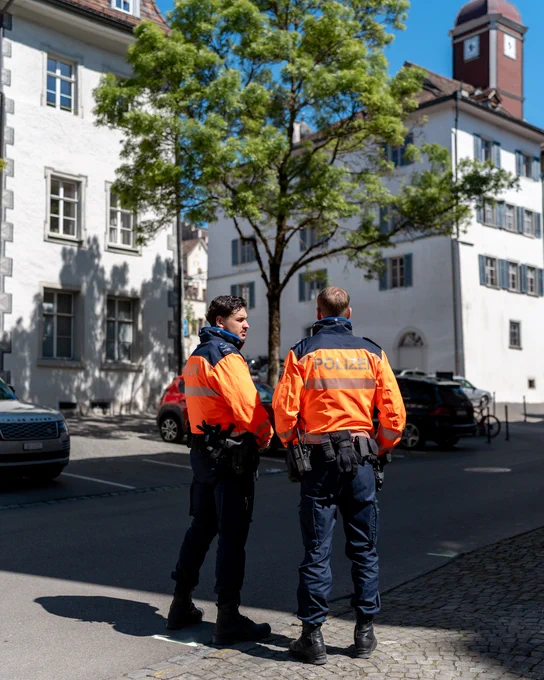Zwei Polizeibeamte in orangefarbenen Warnwesten stehen auf einer Straße zwischen Gebäuden mit Bäumen.