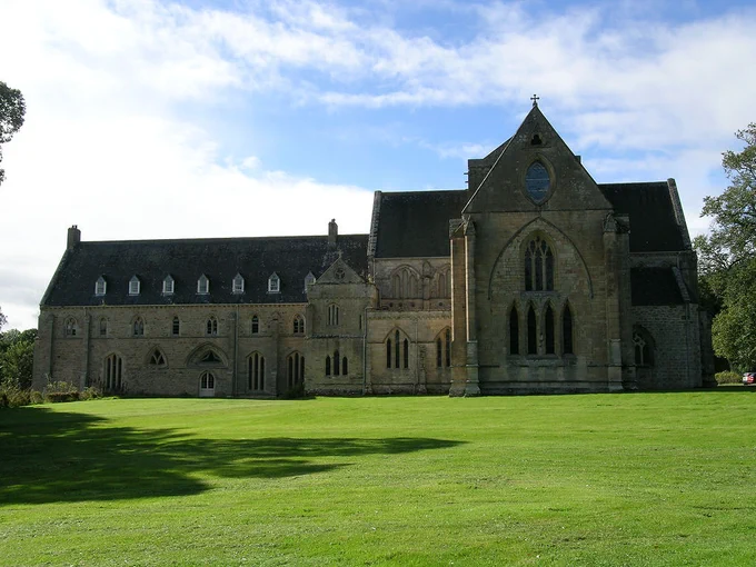 Pluscarden Abbey. The image shows the abbey stretching across the centre of the frame with mown grass in the foreground. The end of the abbey church is on the right and the rest of the buildings are to its left. The scene is in sunlight but the abbey is in its own shadow.