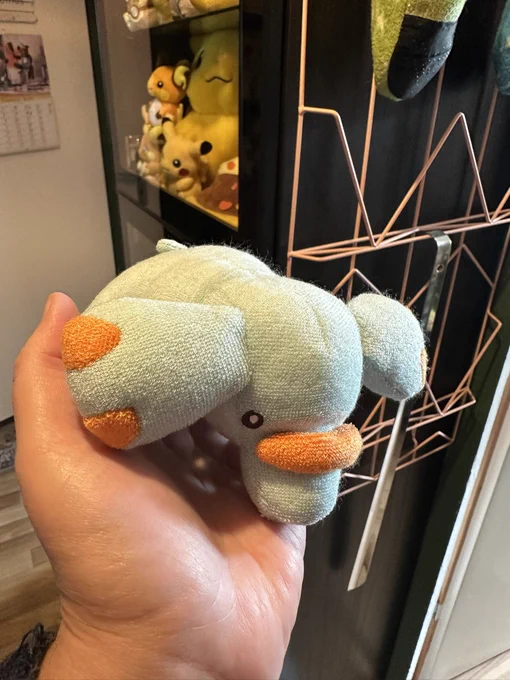 Eine Nahaufnahme einer menschlichen Hand, die ein kleines, hellblaues Kuscheltier des Pokémons Phanpy hält. Das Stofftier hat eine gestrickte Textur, große abstehende Ohren und orangefarbene Akzente an den Zehen und auf dem Rüssel. Im verschwommenen Hintergrund sind ein Regal mit weiteren Pokémon-Plüschtieren (Pikachu) und ein rosanes Metallgitter zu erkennen.