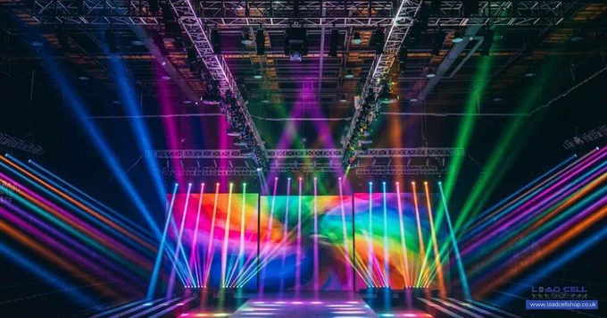 A vibrant concert stage set-up with colourful spotlights and beams shining in various directions, illuminating a large LED screen displaying swirling rainbow patterns and supported by advanced stage rigging with integrated load cells.