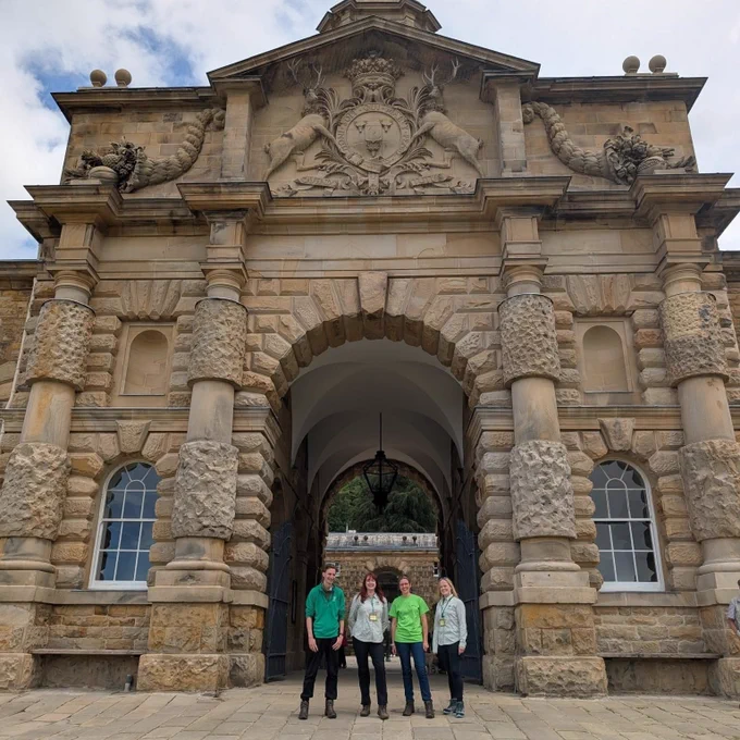 A group of people stands in front of the grand entrance of Chatsworth House, featuring an ornate stone archway. The arched entrance is detailed with intricate carvings. The sky is partly cloudy.