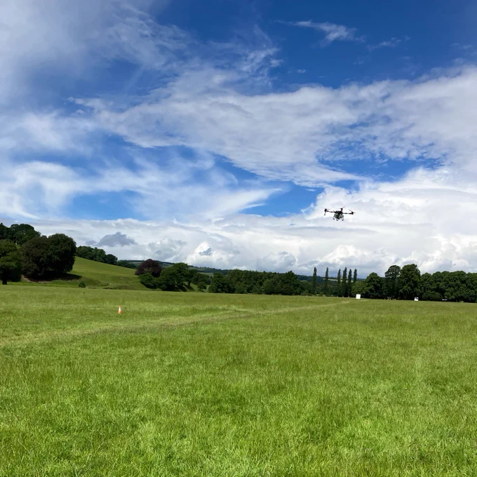 A drone flies over a lush, green field under a bright blue sky with scattered clouds. A single orange cone is visible in the field. Trees and rolling hills line the horizon.