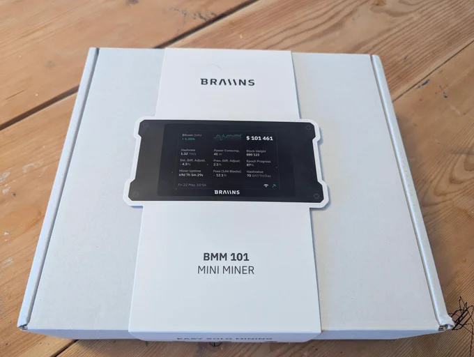 A box containing a Braiin BMM 101 Mini Miner, featuring a digital display showing various mining statistics. The packaging rests on a wooden surface.