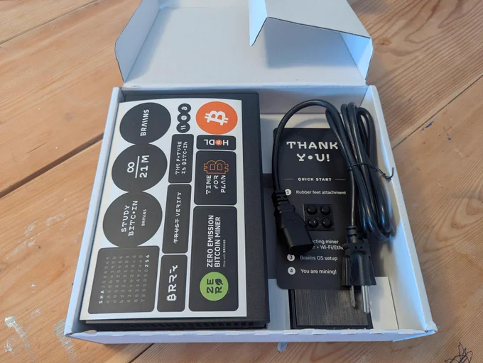 An open box containing Bitcoin mining equipment includes stickers, a power cable, and setup instructions. The minimalist design and tech-focused theme convey a modern and innovative feel.